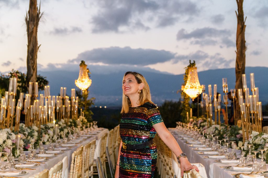 In questa foto si vede uno degli allestimenti de Le Farfalle in Fiore - Angela Crupi: una lunga tavolata allestita all’esterno tra le luci dorate dei candelabri e lampadari sospesi. La wedding planner, in abito arcobaleno di paillettes, sorride al tramonto con le montagne sullo sfondo.