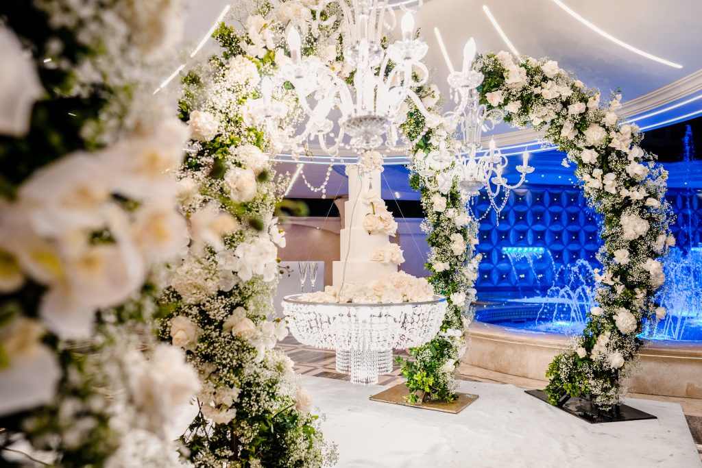 In questa foto si vede una delle tendenze allestimenti location matrimonio 2026: una torta nuziale bianca a più piani, sospesa su un elegante lampadario di cristallo, circondata da archi floreali bianchi e verdi. Il contesto è sofisticato, con illuminazione blu in sottofondo e una fontana d'acqua che crea un'atmosfera scenografica e lussuosa.