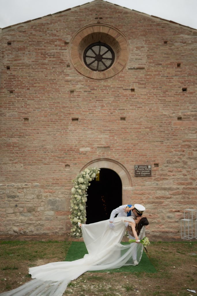 In questa foto si vede una delle caratteristiche dei matrimoni Gen Z, con gli sposi che si baciano davanti a una chiesa in mattoni in una posa dinamica e spontanea, lontana dalla formalità tradizionale.