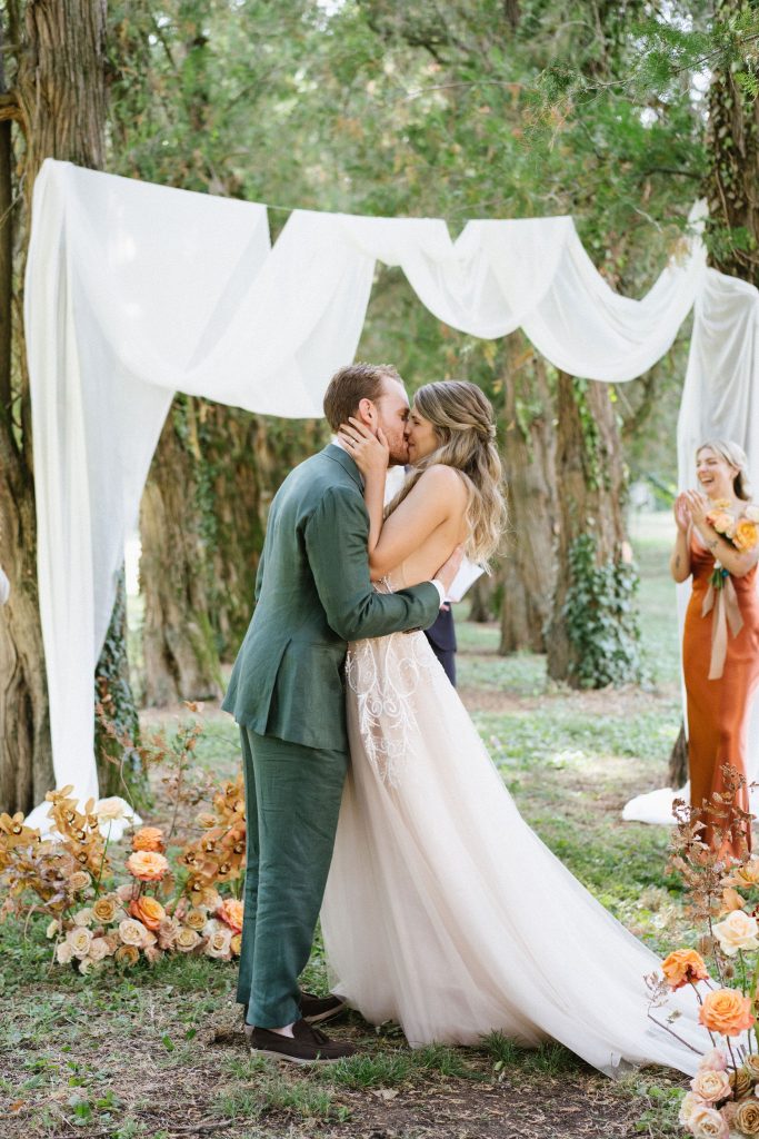 In questa foto di vede una delle caratteristiche dei matrimoni Gen Z, attraverso un bacio tra gli sposi sotto un arco di tessuti leggeri immerso nel verde, con allestimenti naturali e atmosfera intima.