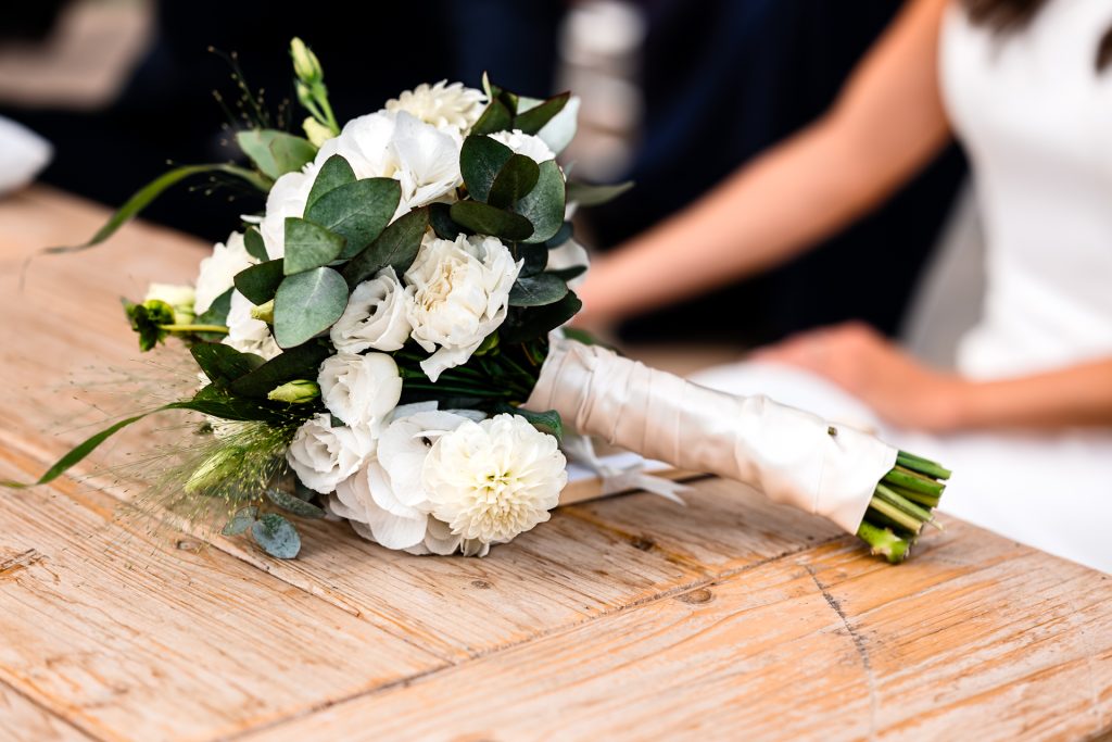 In questa foto si vede uno degli ambienti dedicati al ricevimento matrimonio al Phi Hotel Piajo: un bouquet nuziale adagiato su un tavolo in legno naturale. La composizione è elegante e romantica, con fiori bianchi e verde eucalipto, perfettamente avvolti da un nastro bianco in raso. Sullo sfondo, sfocata, si intravede la sposa con un abito bianco.