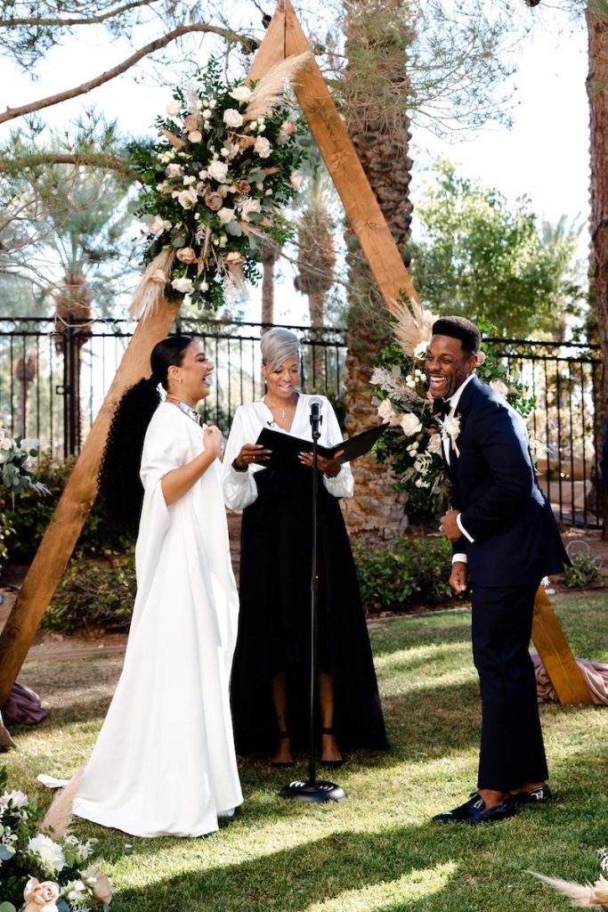 In questa foto due sposi ridono davanti ad un altare di legno di forma triangolare decorato con fiori e spighe durante il loro matrimonio civile a Las Vegas