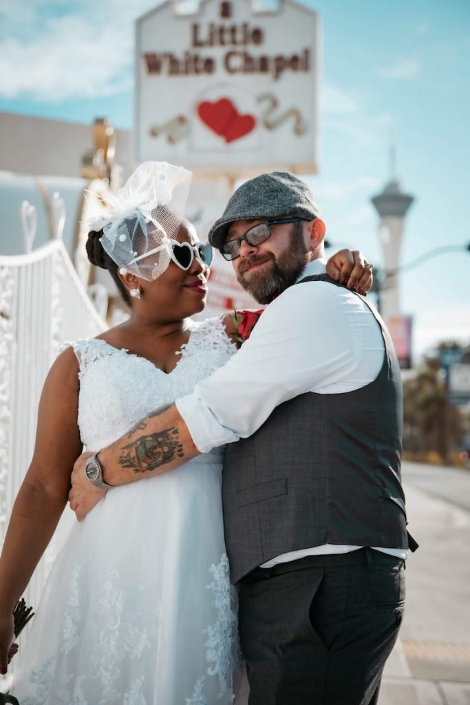 In questa foto due sposi sorridenti e abbracciati davanti all'insegna della Little White Chapel