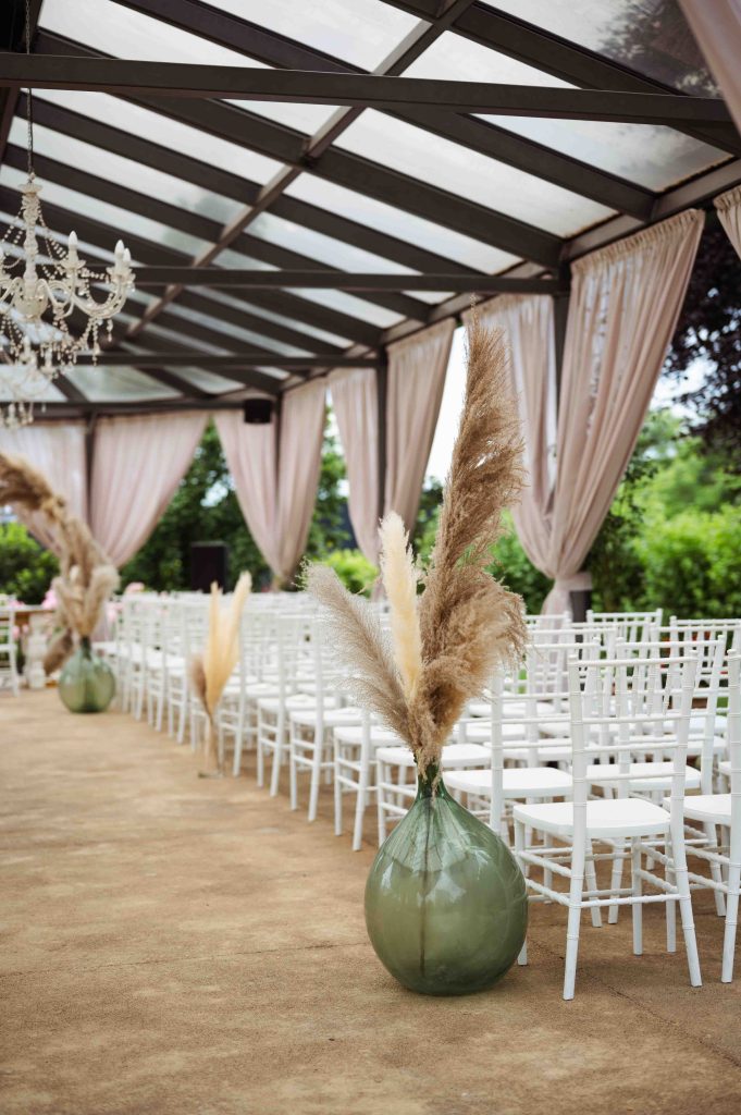 In questa foto si vede uno degli ambienti dedicati al ricevimento matrimonio al Phi Hotel Piajo: un’area cerimonia coperta da una struttura in ferro e vetro, decorata con pampas in grandi vasi di vetro verde. Le sedie bianche sono disposte ordinatamente su un tappeto color sabbia, pronte ad accogliere gli ospiti.