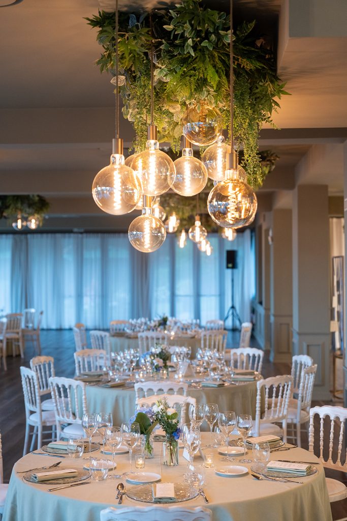 In questa foto si vede uno degli ambienti dedicati al ricevimento matrimonio al Phi Hotel Piajo: un’elegante sala interna allestita per il banchetto, con tavoli tondi ben apparecchiati, sedie chiare e una cascata di lampade tonde dal soffitto decorate con verde sospeso.