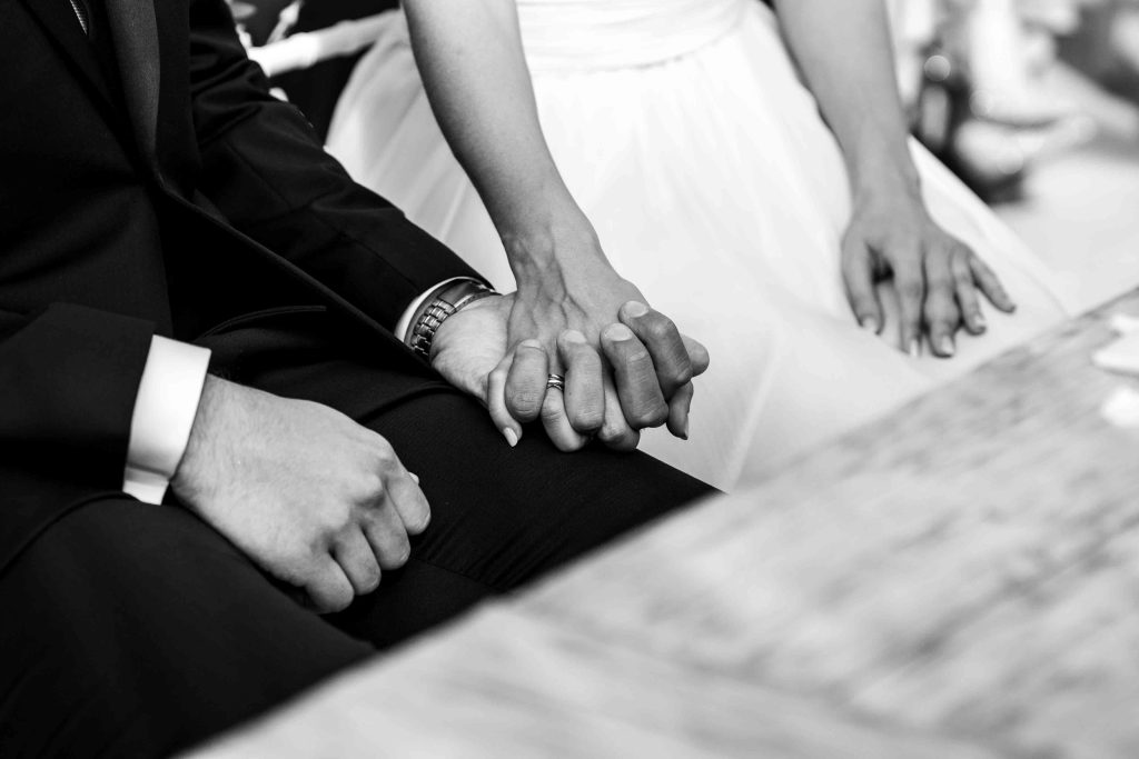 In questa foto si vede un momento intimo catturato in bianco e nero di un matrimonio al Phi Hotel Piajo che mostra gli sposi mentre si tengono per mano, seduti una accanto all’altro. Il gesto affettuoso è enfatizzato dal contrasto tra l'abito scuro dello sposo e il bianco del vestito della sposa.