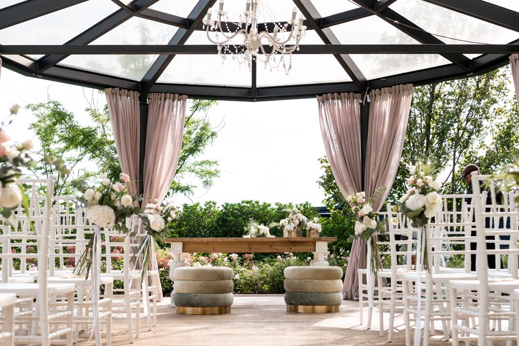 In questa foto si vede uno degli ambienti dedicati al ricevimento matrimonio al Phi Hotel Piajo: una raffinata area cerimonia all’aperto allestita sotto un gazebo trasparente con struttura in ferro nero e tende cipria. Le sedie bianche sono disposte ordinatamente su entrambi i lati del corridoio centrale, con delicati fiori bianchi e rosa applicati sui lati. Al centro, due pouf rotondi imbottiti e un tavolo in legno chiaro creano la scenografia perfetta per il momento del sì.