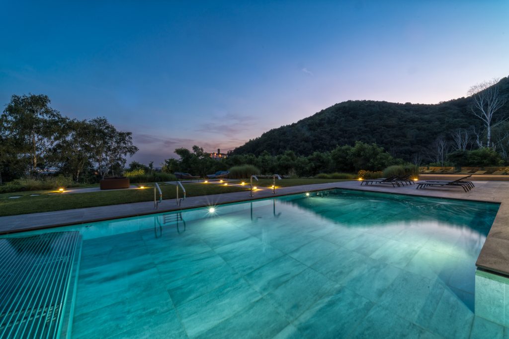 In questa foto si vede uno degli ambienti dedicati al matrimonio al Phi Hotel Piajo: la piscina esterna al tramonto, circondata dal verde della natura e da sdraio scure disposte lungo i bordi. L’acqua azzurra riflette la luce tenue del cielo serale, creando un effetto suggestivo e rilassante. Sullo sfondo si intravede una collina boscosa, che completa il paesaggio.
