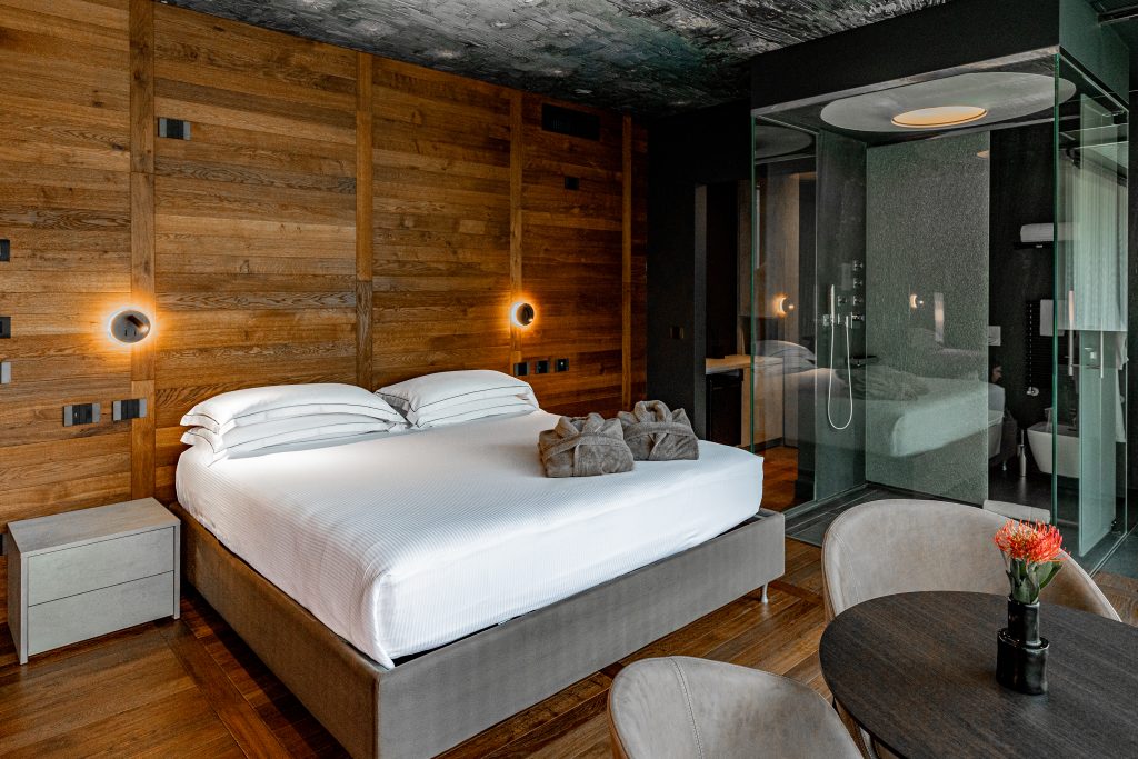 In questa foto si vede uno degli ambienti dedicati al matrimonio al Phi Hotel Piajo: una camera matrimoniale di design, con pareti in legno, dettagli moderni e una doccia a vista racchiusa in vetro. Il letto è ampio, vestito di bianco, e due accappatoi piegati sono adagiati sopra.