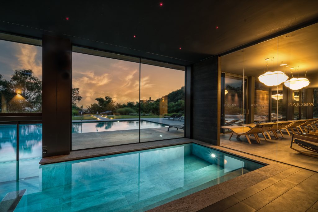 In questa foto si vede uno degli ambienti dedicati al matrimonio al Phi Hotel Piajo: la piscina interna riscaldata, situata accanto a una grande vetrata che si affaccia sull’esterno. All’esterno si intravede la piscina scoperta illuminata, mentre all’interno l’ambiente è caldo, avvolgente e raffinato, con lettini in legno chiaro e luci soffuse che valorizzano lo spazio relax.