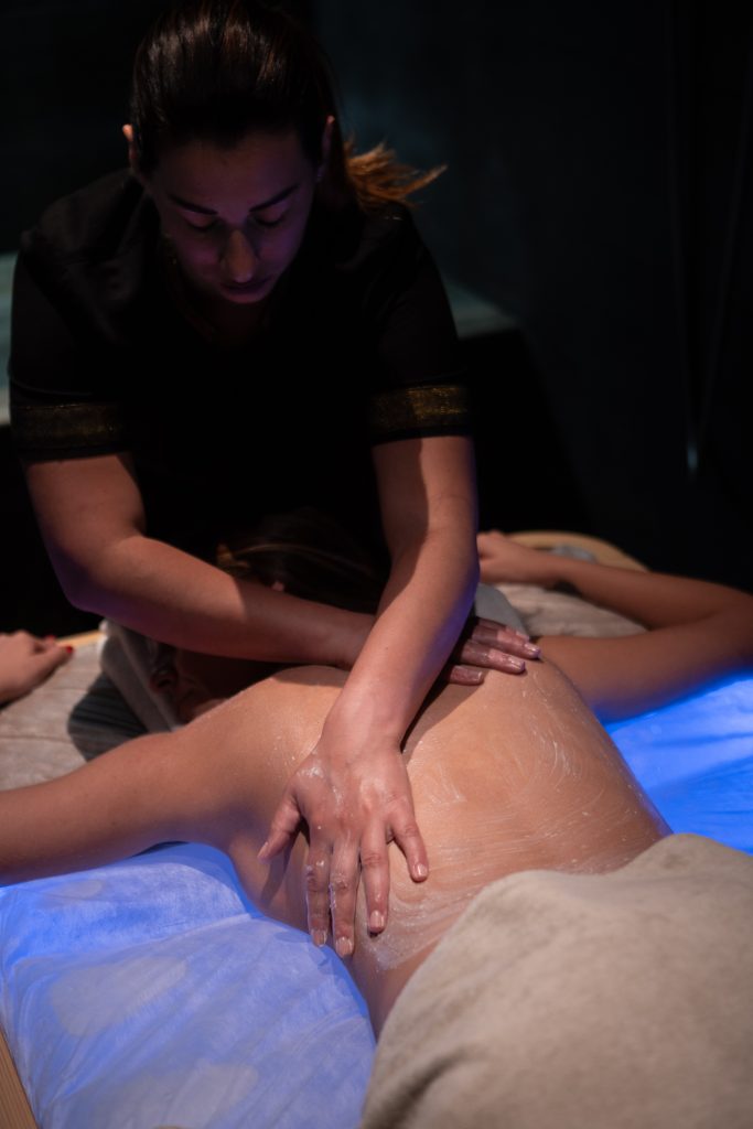 In questa foto si vede uno degli ambienti dedicati al ricevimento matrimonio al Phi Hotel Piajo: la zona benessere della spa, dove una professionista sta eseguendo un massaggio su schiena e spalle. La scena, avvolta da luci soffuse e atmosfera rilassante, trasmette cura e attenzione ai dettagli. La pelle della cliente è cosparsa di crema, e le mani della massaggiatrice si muovono con precisione e delicatezza.