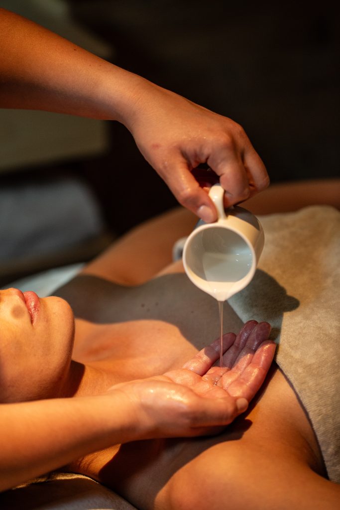 In questa foto si vede uno degli ambienti dedicati al ricevimento matrimonio al Phi Hotel Piajo: un momento di benessere in spa, in cui un’operatrice versa un olio o un latte idratante sulle mani prima di procedere con il trattamento. In primo piano si nota la pelle luminosa della cliente, sdraiata sul lettino, e l’atmosfera rilassante creata dalla luce calda e soffusa.