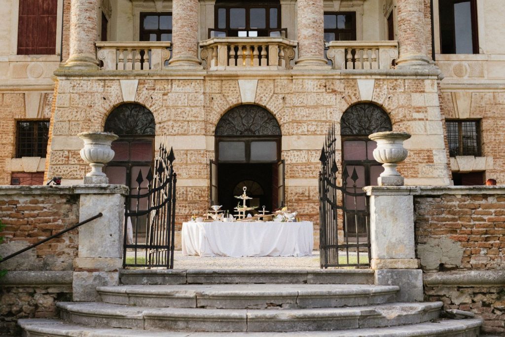 In questa foto si vede uno dei momenti di un matrimonio intimo Villa Francanzan Piovene: all’ingresso monumentale della villa, una mise en place bianca con alzate e vassoi è pronta per il servizio dolci o aperitivo. L’inquadratura valorizza la pietra viva e la maestosità architettonica del luogo.