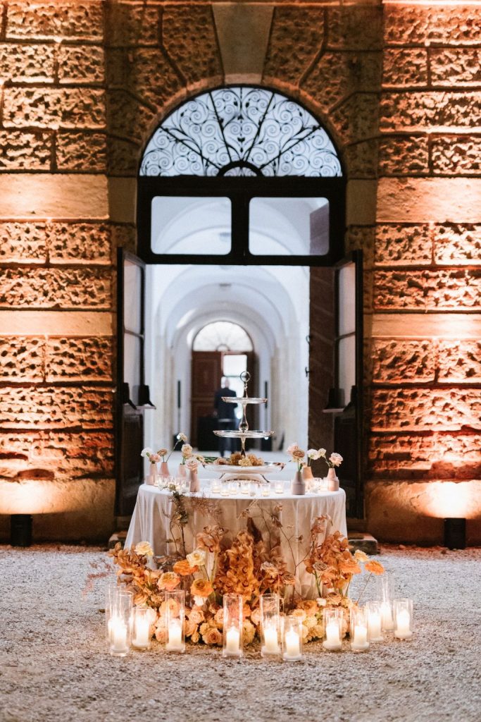 In questa foto si vede uno dei momenti di un matrimonio intimo Villa Francanzan Piovene: un tavolo rotondo allestito nella corte esterna, illuminato da decine di candele in cilindri di vetro. I fiori, nelle sfumature dell’arancio bruciato e del rosa cipria, decorano la base del tavolo, creando un’atmosfera calda e accogliente per il dessert o il taglio torta.