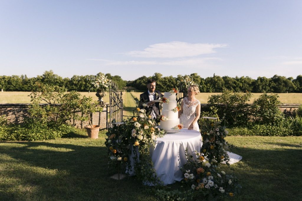 In questa foto si vede uno dei momenti di un matrimonio intimo Villa Francanzan Piovene: gli sposi si preparano a tagliare una torta nuziale monumentale, circondata da una scenografia floreale che richiama i colori del giardino. Sullo sfondo, un viale alberato che si perde all’orizzonte completa la scena bucolica.
