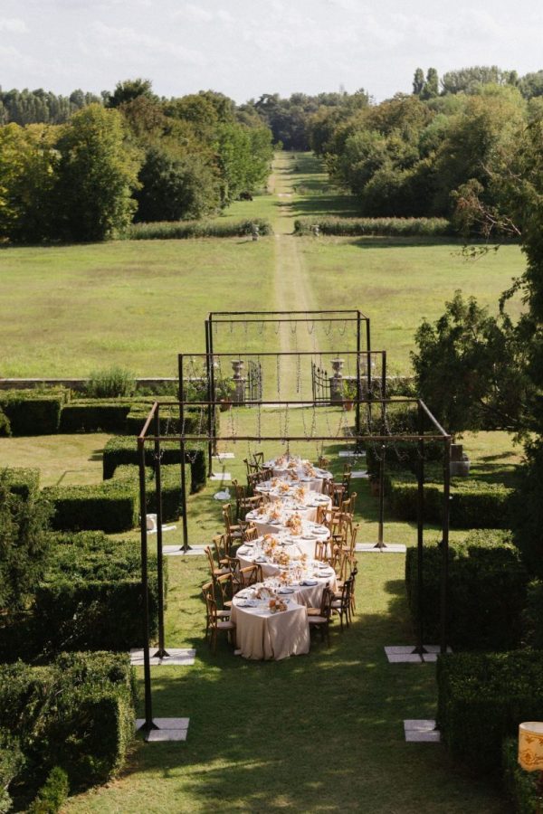 In questa foto si vede uno dei momenti di un matrimonio intimo Villa Francanzan Piovene: una prospettiva centrale mostra la simmetria perfetta dell’allestimento outdoor, con i tavoli disposti lungo un viale erboso, sotto una struttura metallica pronta ad accogliere luci o decorazioni sospese.