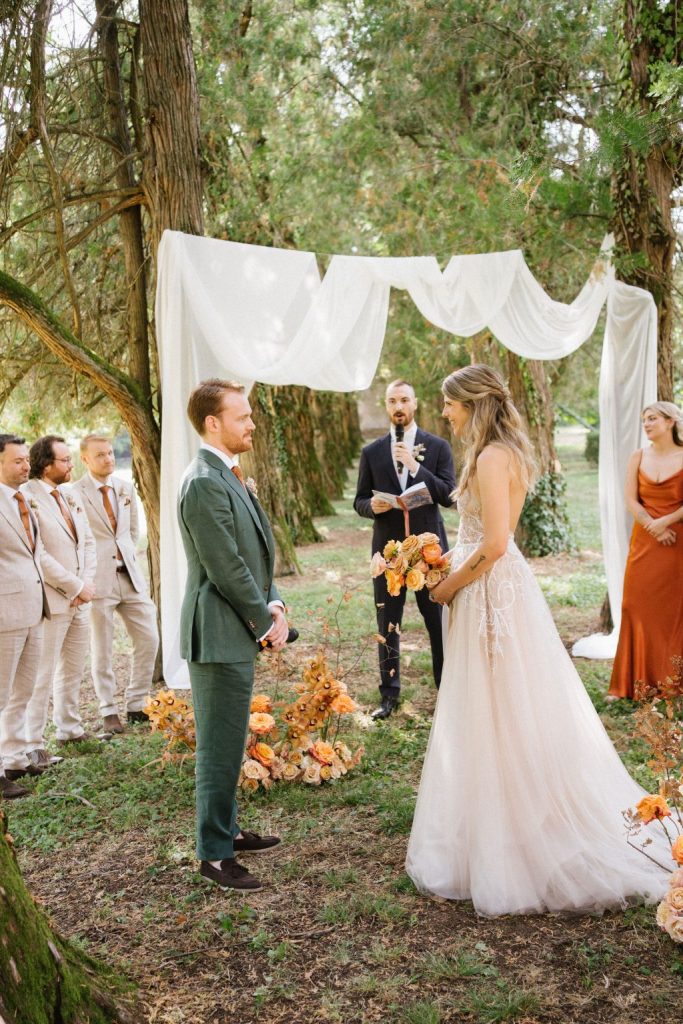 In questa foto si vede uno dei momenti di un matrimonio intimo Villa Francanzan Piovene: la cerimonia si svolge in un boschetto, con l’altare decorato da drappi bianchi e fiori arancio. Gli sposi si scambiano le promesse circondati da testimoni vestiti in toni neutri e caldi.