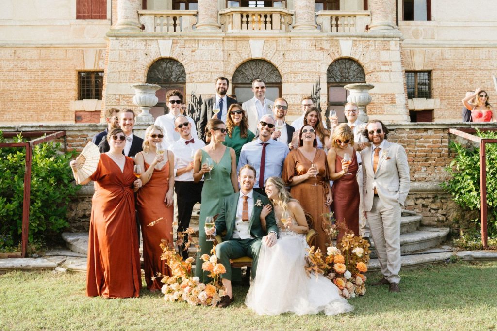 In questa foto si vede uno dei momenti di un matrimonio intimo Villa Francanzan Piovene: un ritratto di gruppo spensierato raccoglie amici e testimoni attorno agli sposi. I colori caldi dei loro outfit e la composizione floreale arancio e pesca evocano un mood elegante ma informale.
