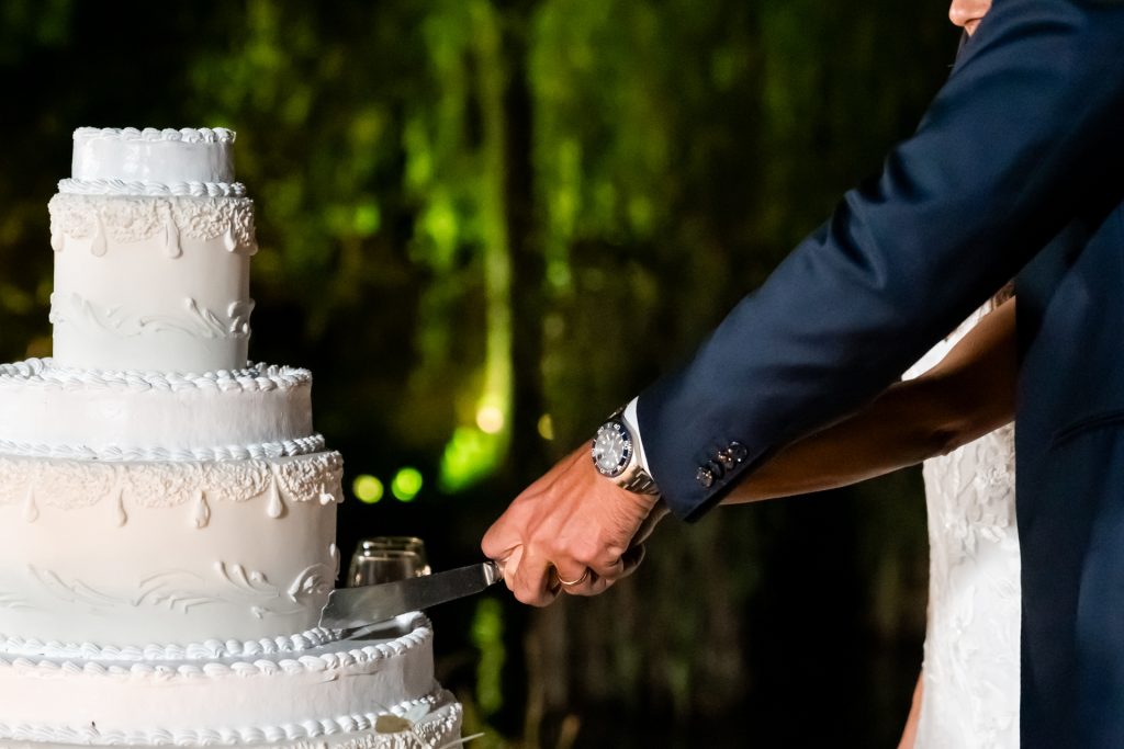 In questa foto si vede uno degli ambienti dedicati al ricevimento matrimonio al Phi Hotel Piajo: un momento intimo e simbolico in cui gli sposi tagliano insieme la torta nuziale. La torta, imponente e a più piani, è bianca con eleganti decori in rilievo. In primo piano si vede la mano dello sposo che afferra il coltello, indossando un orologio dal cinturino metallico, mentre la sposa lo accompagna con un gesto complice.