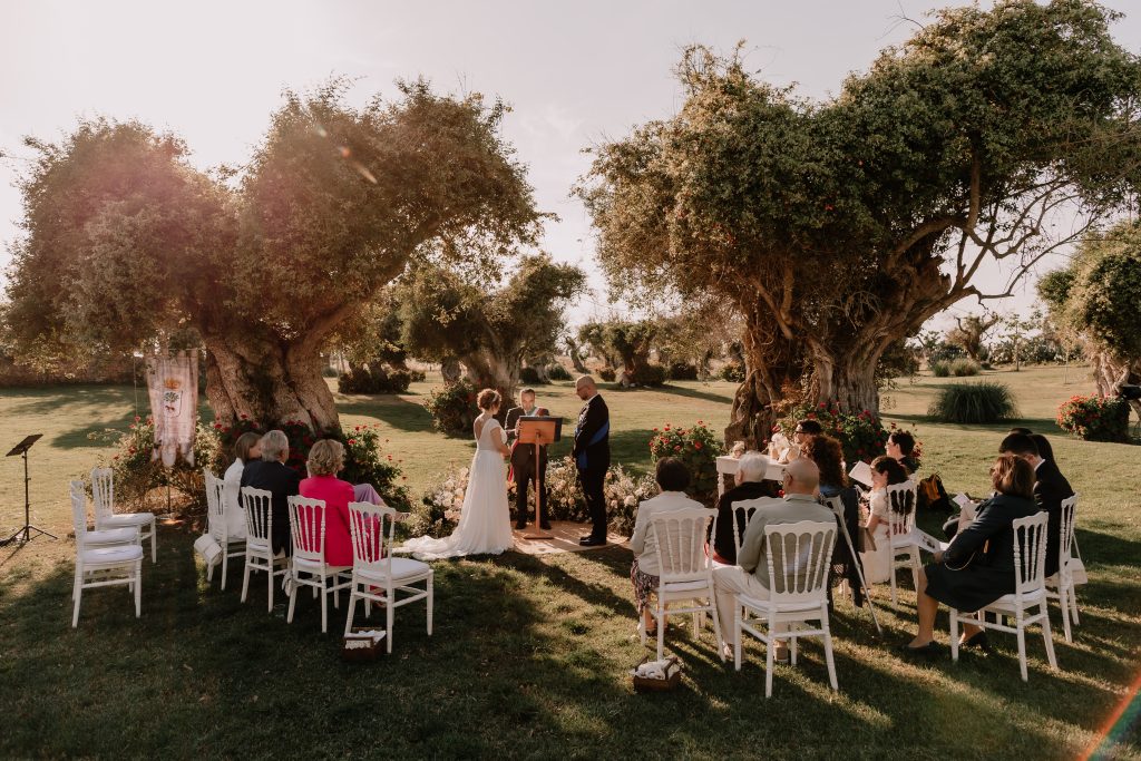 In questa foto si vede uno degli spazi di un matrimonio a a Tenuta Monacelli, la cerimonia civile all’aperto sotto ulivi secolari, con sedie bianche disposte ordinatamente e gli sposi al centro del prato.