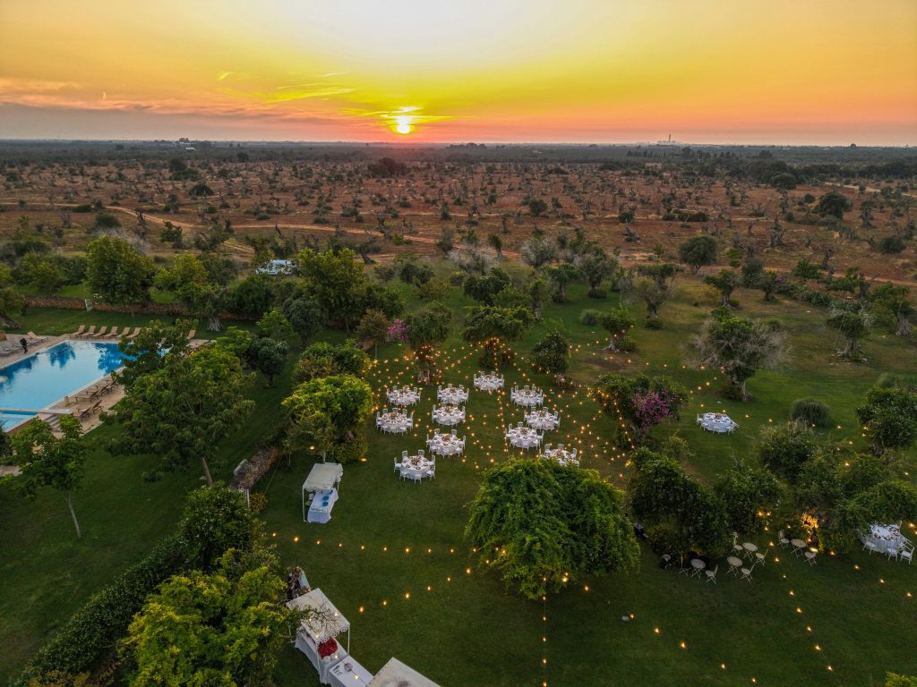 In questa foto si vede uno degli spazi di un matrimonio a a Tenuta Monacelli, una veduta panoramica dall’alto del ricevimento serale nel giardino, con tavoli rotondi illuminati da luci calde e la campagna pugliese al tramonto.