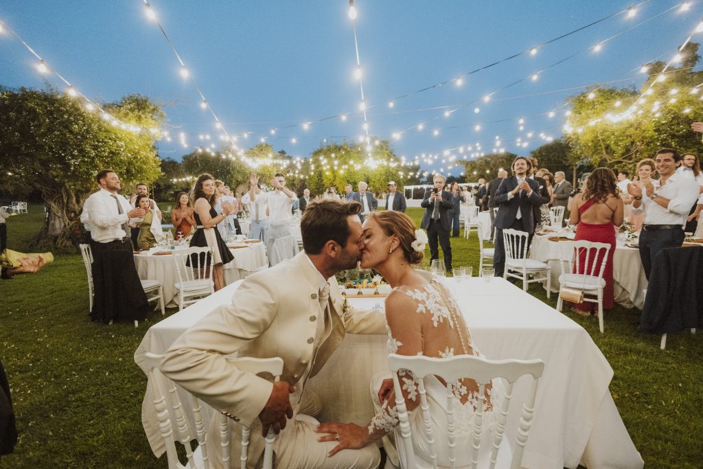 In questa foto si vede uno degli spazi di un matrimonio a a Tenuta Monacelli, gli sposi seduti al tavolo d’onore che si baciano all’aperto, circondati dagli invitati e da luci sospese tra gli alberi.