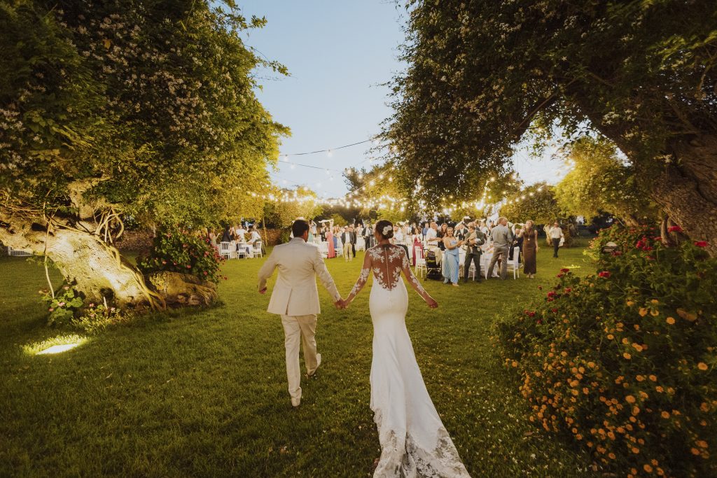 In questa foto si vede uno degli spazi di un matrimonio a a Tenuta Monacelli, gli sposi che camminano mano nella mano nel giardino illuminato da luci sospese, mentre gli invitati li attendono sullo sfondo.