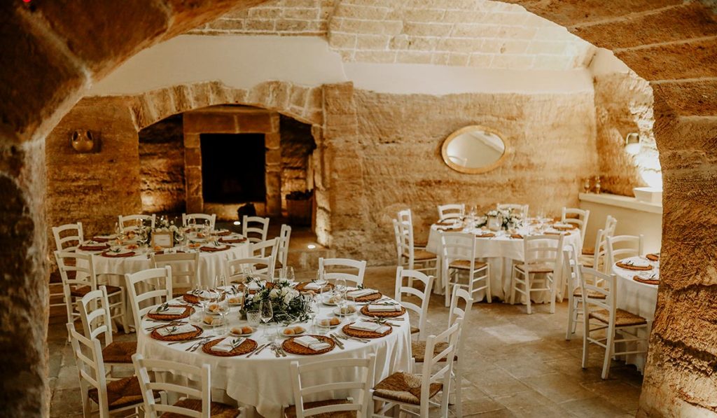 In questa foto si vede uno degli spazi di un matrimonio a a Tenuta Monacelli, una sala interna in pietra con tavoli rotondi apparecchiati, sedie bianche e un’atmosfera calda e raccolta.