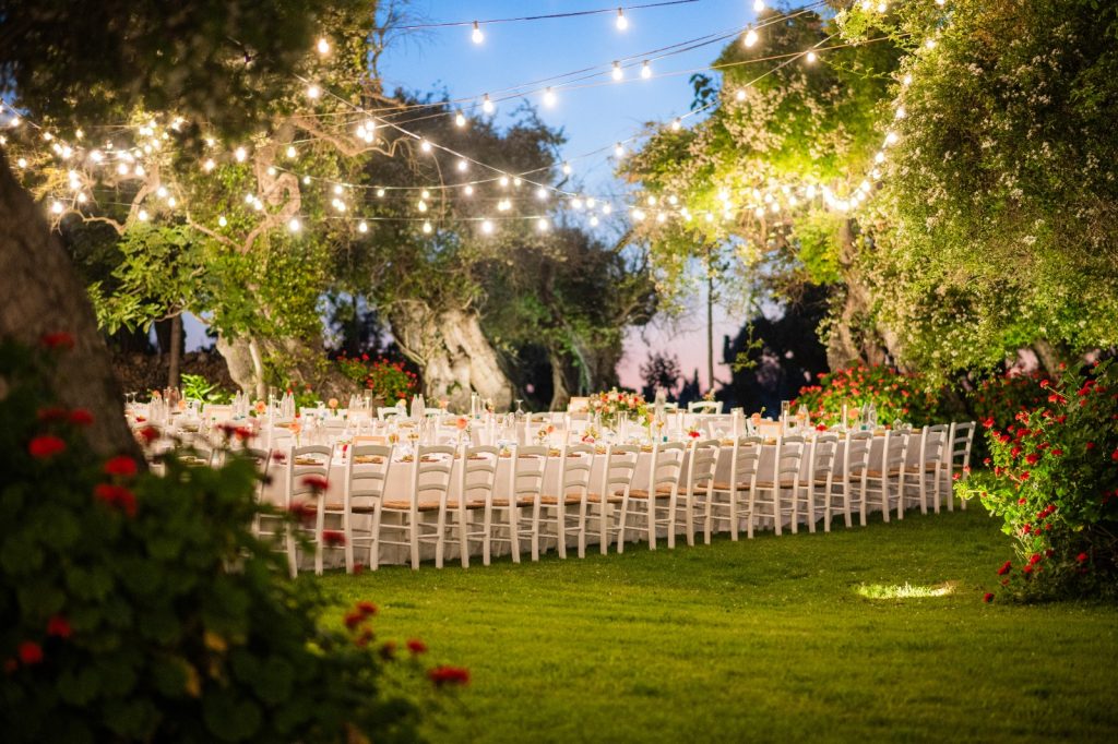 In questa foto si vede uno degli spazi di un matrimonio a a Tenuta Monacelli, una lunga tavolata imperiale all’aperto, illuminata da luci sospese tra gli alberi, immersa nel verde del giardino al tramonto.