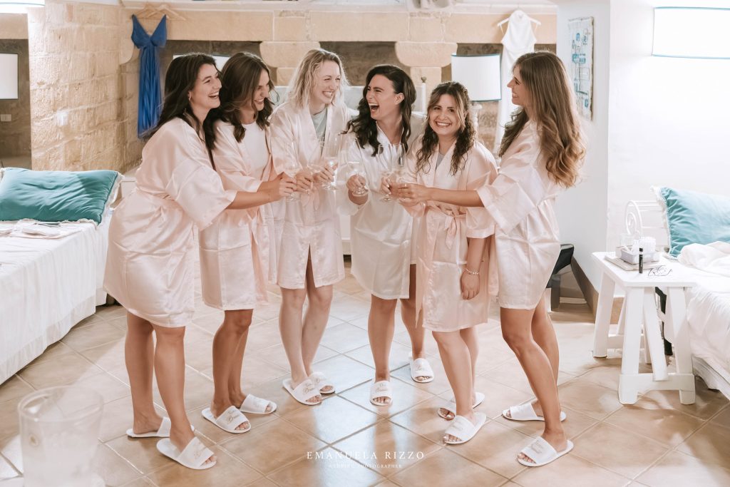 In questa foto si vede uno degli spazi di un matrimonio a a Tenuta Monacelli, la preparazione della sposa con le amiche, tutte in vestaglie chiare, mentre brindano sorridendo in una luminosa stanza in pietra.
