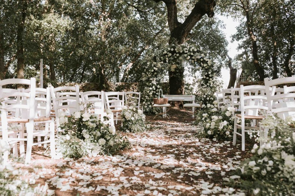 In questa foto si vede uno degli spazi di un matrimonio a a Tenuta Monacelli, l’allestimento della cerimonia tra gli alberi, con sedie bianche, petali a terra e un arco floreale in tonalità chiare.