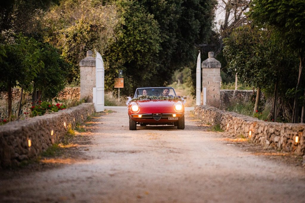In questa foto si vede uno degli spazi di un matrimonio a a Tenuta Monacelli, l’arrivo degli sposi su un’auto d’epoca rossa che attraversa il viale d’ingresso della tenuta incorniciato da muretti in pietra.