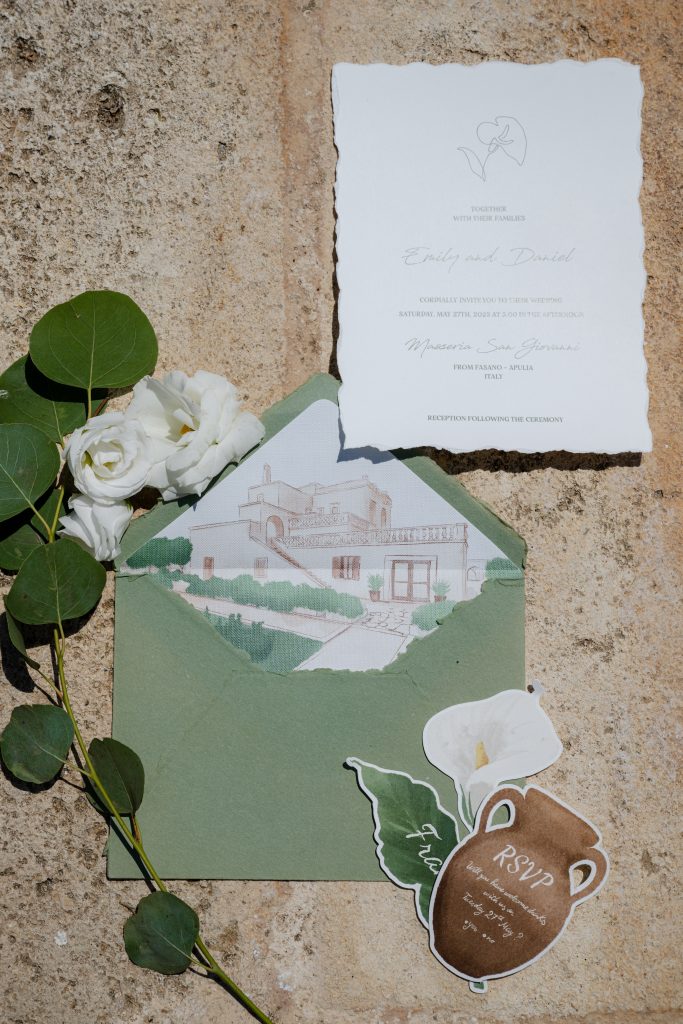 In questa foto si vede una delle tendenze partecipazioni matrimonio 2026: una suite raffinata in stile rustico-elegante con invito stampato su carta artigianale dai bordi sfrangiati, accompagnato da una busta verde salvia che svela all’interno un’illustrazione acquerellata della location. Completano il set un cartoncino RSVP a forma di anfora in terracotta, una calla dipinta e una composizione naturale con fiori bianchi e rami di eucalipto, il tutto adagiato su una superficie in pietra chiara che richiama l’atmosfera delle masserie pugliesi.