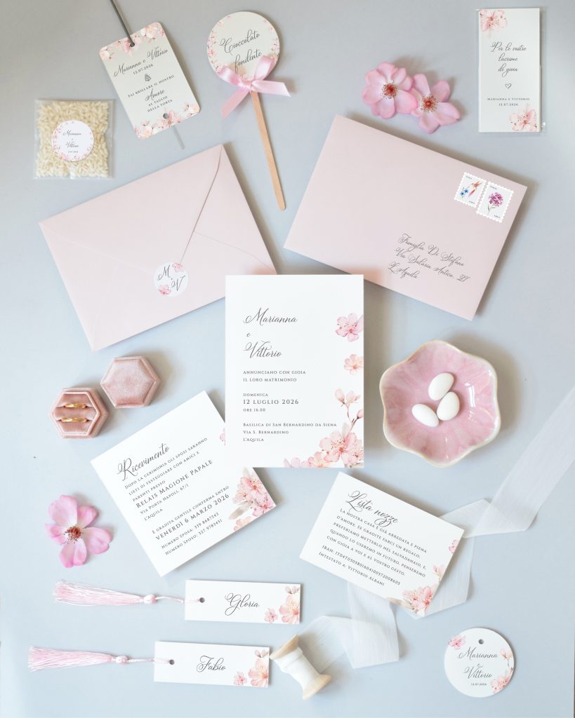 In questa foto si vede una delle tendenze partecipazioni matrimonio 2026: una suite grafica ispirata ai fiori di ciliegio, con inviti, tag, buste e cartoncini in rosa cipria decorati con delicate illustrazioni floreali. Gli elementi coordinati, dalle partecipazioni ai gadget personalizzati, creano un effetto fresco e coerente.