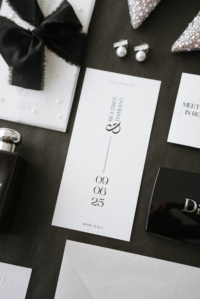 In questa foto si vede una delle tendenze partecipazioni matrimonio 2026: un elegante save the date verticale, stampato su cartoncino bianco con testo nero essenziale. I nomi degli sposi, Beatrice e Damiano, sono scritti in verticale, uniti da un simbolo ampersand, mentre la data del matrimonio (09.06.25) è riportata al centro in caratteri tipografici moderni. Il layout pulito e minimalista è accompagnato da accessori chic, come un profumo nero e dettagli moda in bianco e nero, per un effetto sofisticato e monocromatico.