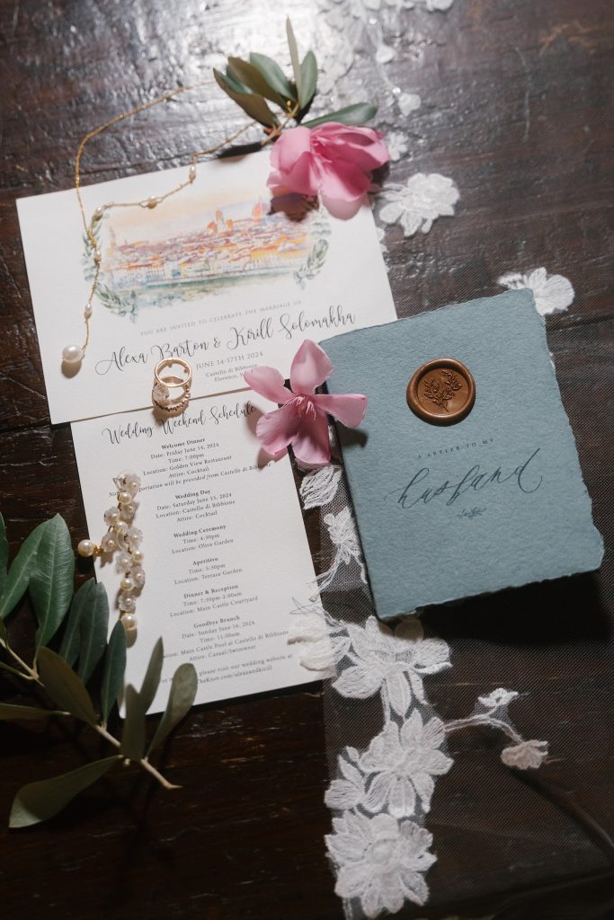 In questa foto si vede una delle tendenze partecipazioni matrimonio 2026: un invito dal tono romantico e artigianale, con acquerello illustrato della location, programma dell’evento in carta avorio e libretto azzurro in carta naturale con sigillo in ceralacca. A completare la scena, fiori freschi, rami d’ulivo e gioielli in perla.