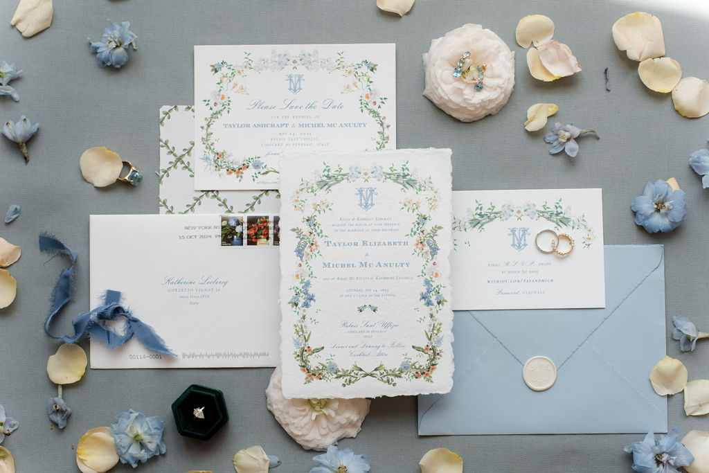 In questa foto si vede una delle tendenze partecipazioni matrimonio 2026: partecipazioni dallo stile romantico e botanico, con cornici di fiori acquerellati in tonalità pastello. I cartoncini sono stampati su carta porosa e accompagnati da buste cerulee, sigilli in cera e francobolli floreali. L’intero set è disposto su un tessuto grigio con petali e accessori in palette, creando un mood poetico e raffinato.