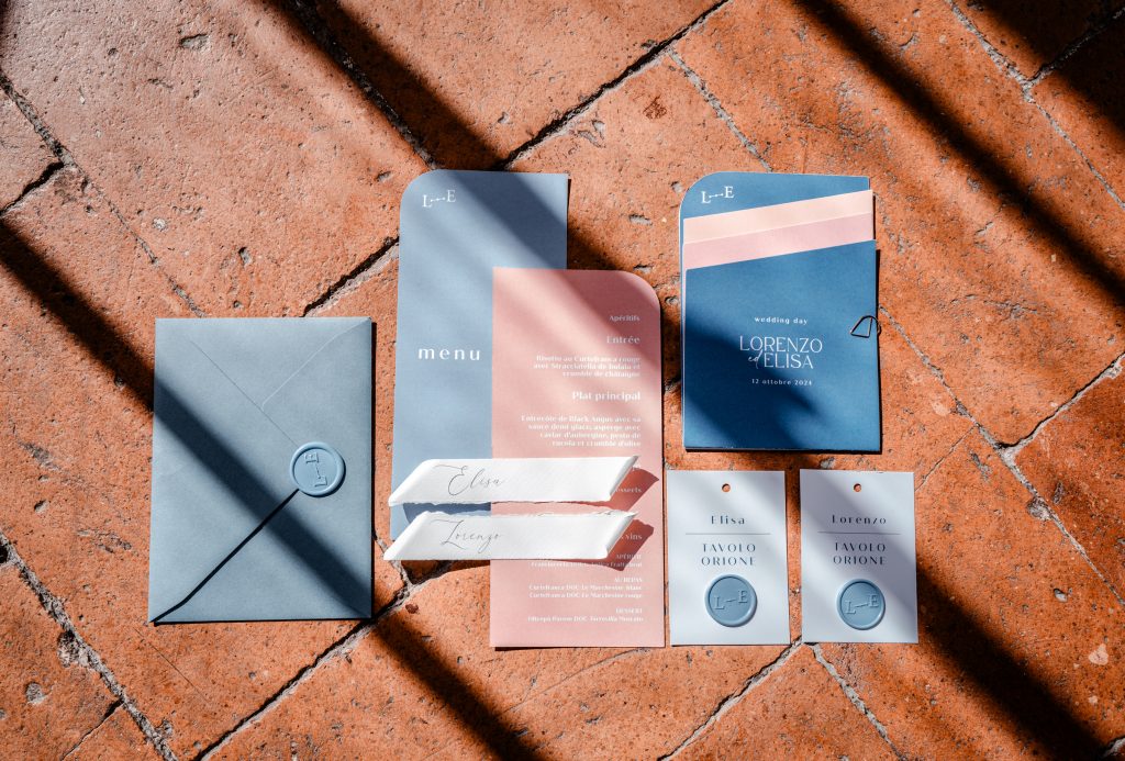 In questa foto si vede una delle tendenze partecipazioni matrimonio 2026: un set grafico moderno, con buste e cartoncini in tonalità azzurro polvere e rosa antico. Il design è geometrico, con angoli arrotondati e font essenziali. Include segnaposto personalizzati, un menù in francese, e sigilli con le iniziali degli sposi. Lo sfondo in cotto rustico crea un contrasto materico con la palette elegante.