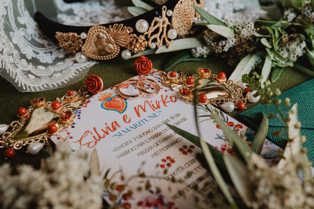 In questa foto si vede una delle tendenze partecipazioni matrimonio 2026: un invito vivace, colorato e simbolico, con motivi sacri, cuori ex voto, elementi dorati e dettagli in rosso corallo. Una scelta scenografica che celebra le radici culturali in chiave contemporanea.