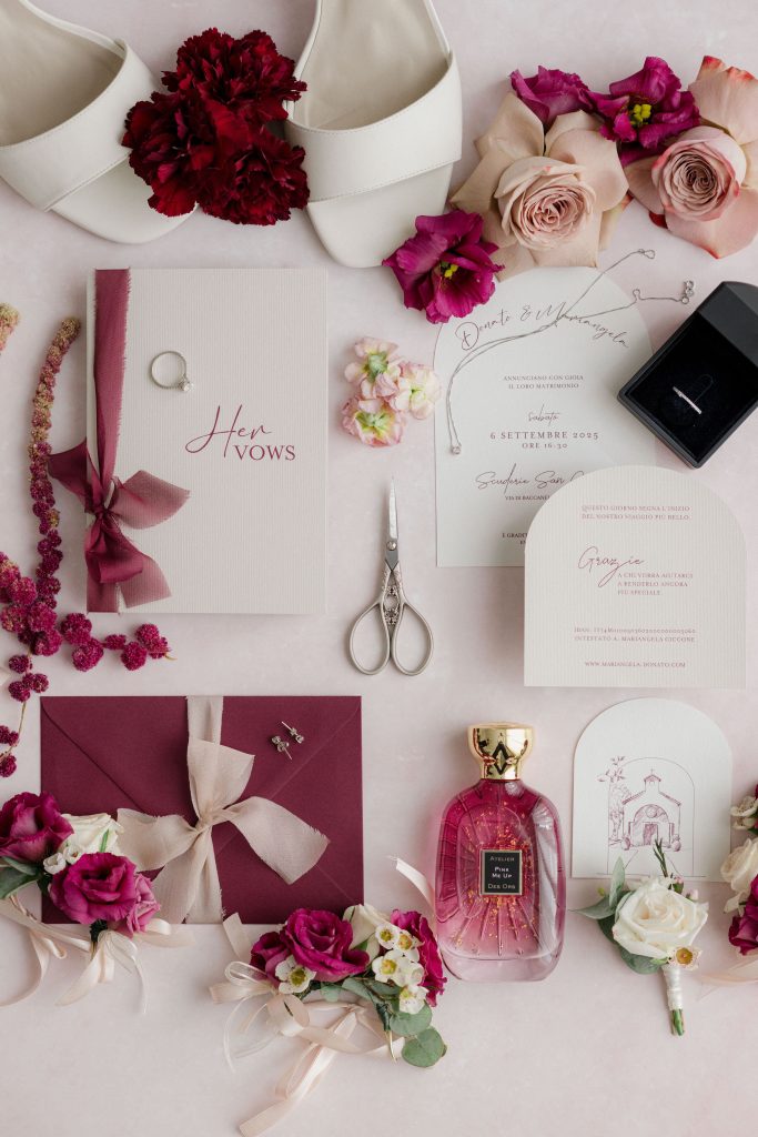 In questa foto si vede una delle tendenze partecipazioni matrimonio 2026: un flat lay floreale in tonalità borgogna e rosa, dove la partecipazione è accompagnata da cartoline “grazie”, libretti dei voti, fedi e profumo. Il colore predominante è il bordeaux, con testi in corsivo elegante. Le partecipazioni hanno forme originali, come l’arco, e si integrano in un tableau visivo ricco di elementi romantici e sensuali.
