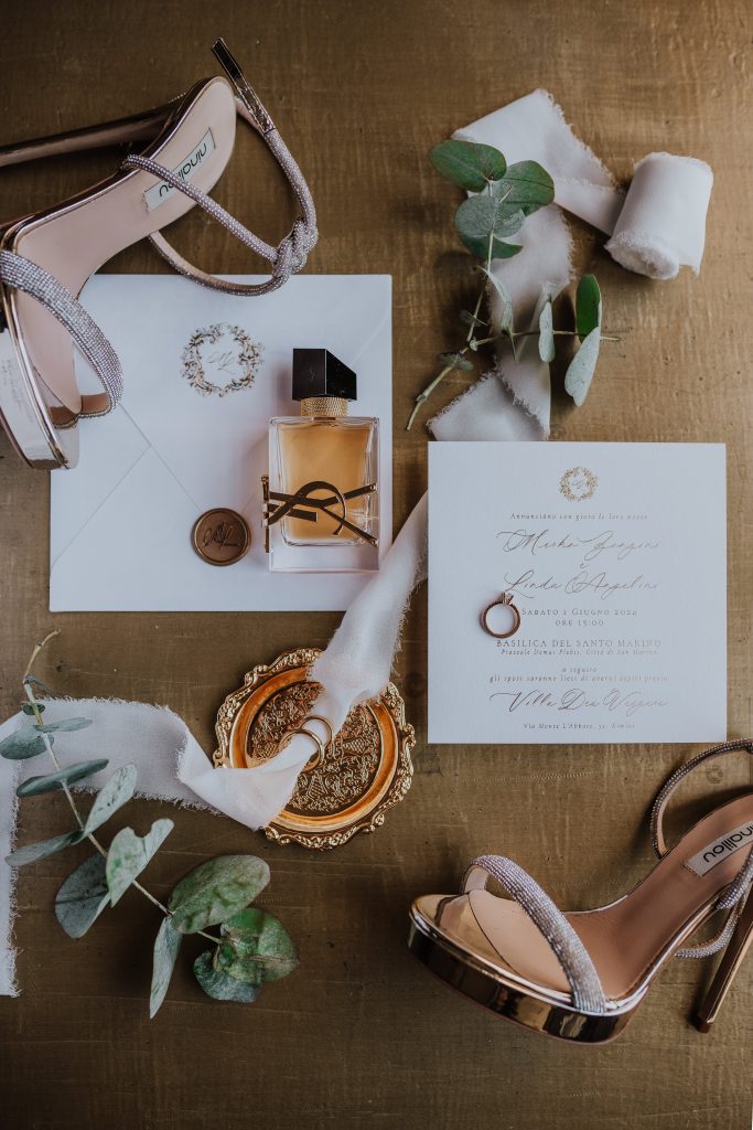 In questa foto si vede una delle tendenze partecipazioni matrimonio 2026: uno stile elegante e glamour, in cui l’invito avorio è arricchito da elementi dorati, nastro di tulle, profumo Yves Saint Laurent e sandali gioiello. Un’allure sofisticata che racconta il tono dell’evento.