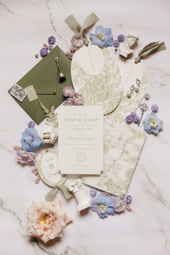 In questa foto si vede una delle tendenze partecipazioni matrimonio 2026: la riscoperta dello stile classico, con inviti color crema, busta verde oliva, grafiche floreali e cartoncini sagomati. Gli accessori – mini statue, chiavi, fiori secchi – arricchiscono l’estetica romantica e senza tempo.