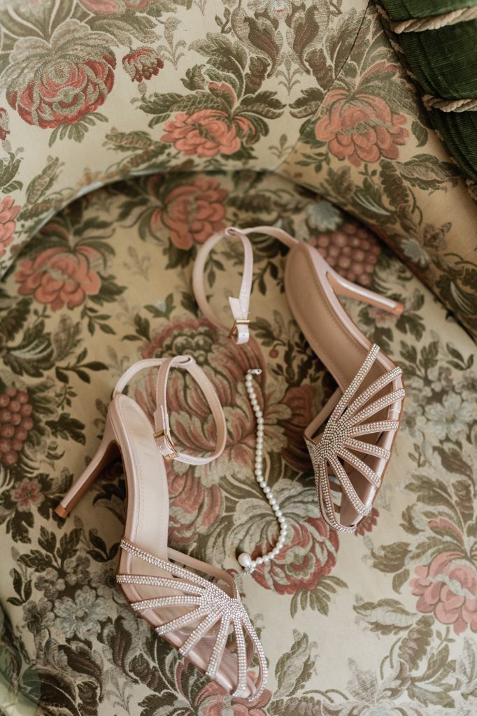 In questa foto si vede una proposta di scarpe da sposa 2026, sandali nude con fasce sottili ricoperte di cristalli, appoggiati su un tessuto floreale insieme a una collana di perle, per un look luminoso e femminile.