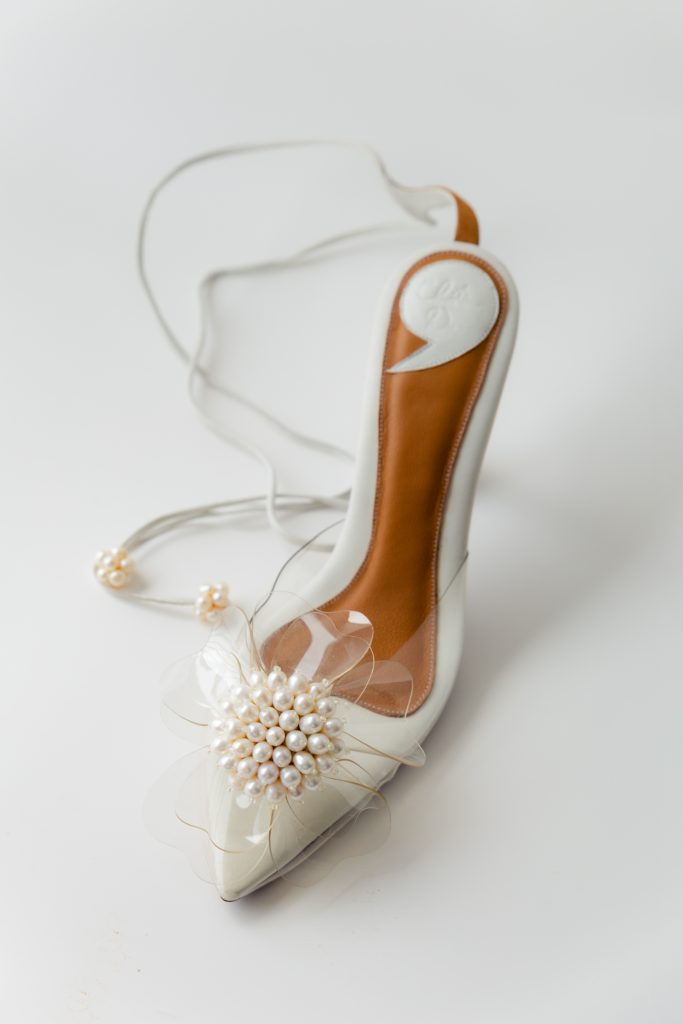 In questa foto si vede una proposta di scarpe da sposa 2026, décolleté bianche con tacco medio e dettaglio decorativo sulla punta, abbinate a un accessorio floreale delicato.