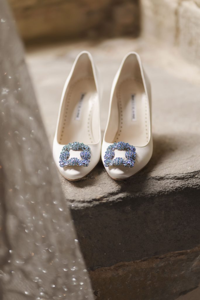 In questa foto si vede una proposta di scarpe da sposa 2026, un paio di décolleté bianche a punta con applicazione gioiello azzurra sulla tomaia, appoggiate su un gradino in pietra accanto a un velo trasparente.