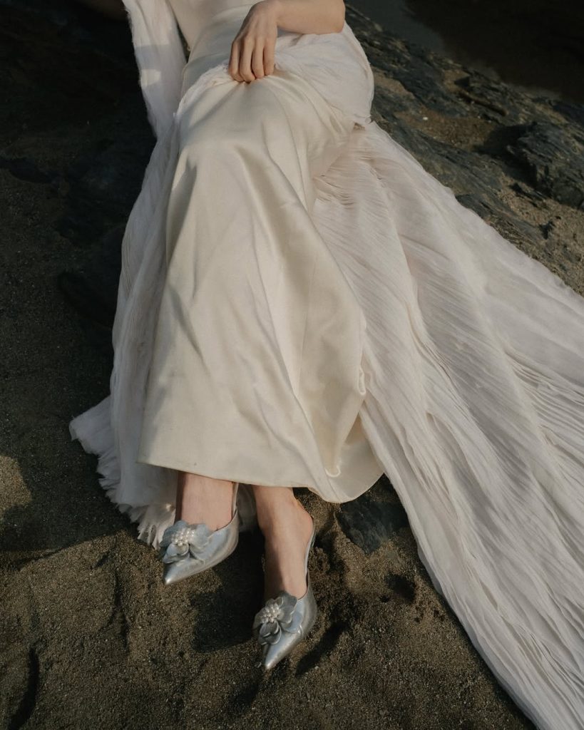 In questa foto si vede una proposta di scarpe da sposa 2026, un paio di ballerine a punta in tonalità argento, con applicazione floreale tridimensionale sul davanti, indossate sotto un abito da sposa chiaro su una spiaggia sabbiosa.