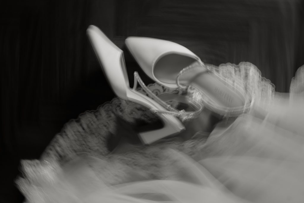 In questa foto si vede una proposta di scarpe da sposa 2026, un paio di décolleté bianche a punta con cinturino gioiello, ritratte in uno scatto artistico in bianco e nero con effetto mosso.