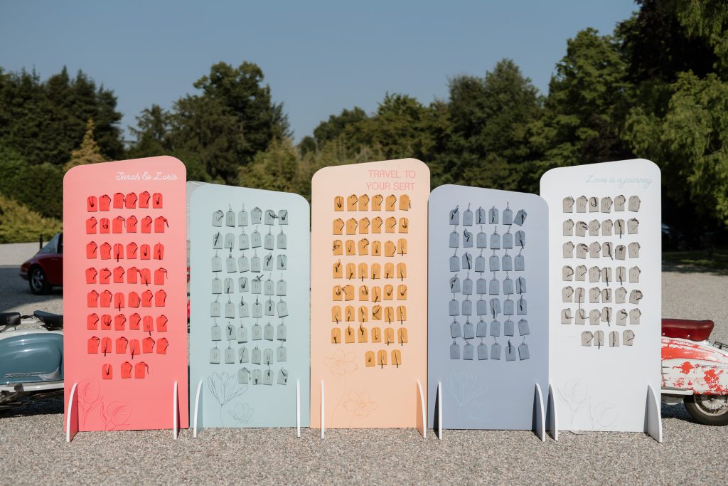 In questa foto si vede una delle tendenze allestimenti location matrimonio 2026: un originale tableau de mariage all’aperto composto da cinque pannelli color pastello, ognuno con etichette simili a tag da valigia. Il design è giocoso e moderno, con scritte come “Travel to your seat” e “Love is a journey”, per un matrimonio a tema viaggio.