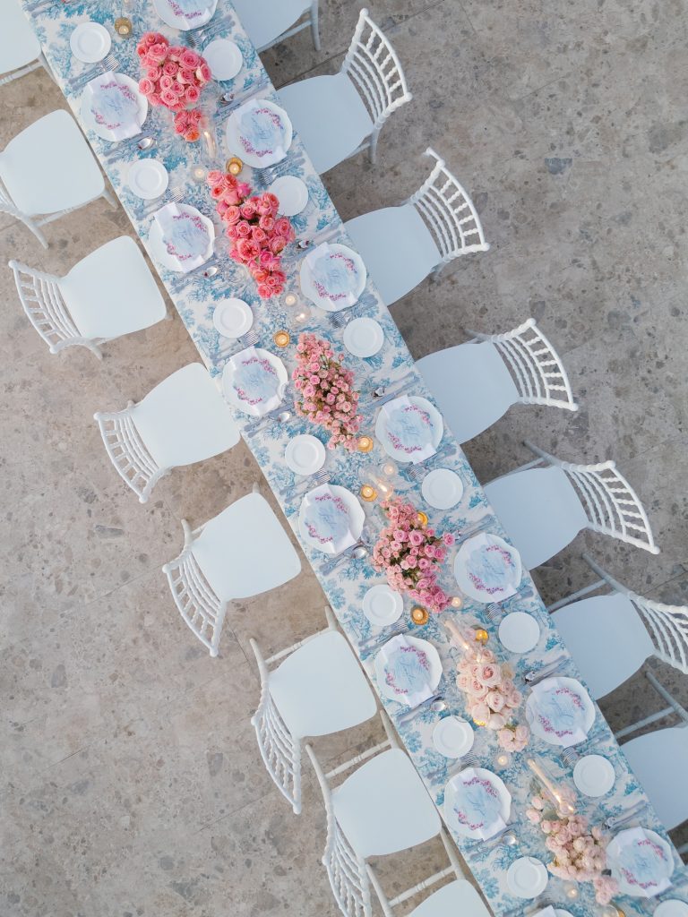 In questa foto si vede una delle tendenze allestimenti location matrimonio 2026: un tavolo da ricevimento ripreso dall’alto, decorato con tovaglie azzurro toile de jouy, piatti decorati e centrotavola di rose rosa disposte a cascata. Le sedie bianche in stile chiavarino completano l’eleganza luminosa della mise en place.