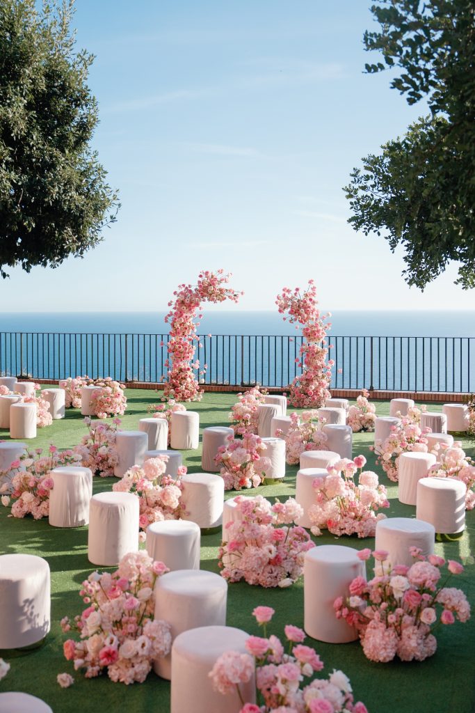 In questa foto si vede una delle tendenze allestimenti location matrimonio 2026: un’area cerimonia all’aperto con sedute rotonde rivestite di bianco, alternate a nuvole di fiori rosa cipria. Due archi floreali speculari incorniciano il punto in cui si svolge la cerimonia, affacciata sul mare e incorniciata da alberi.